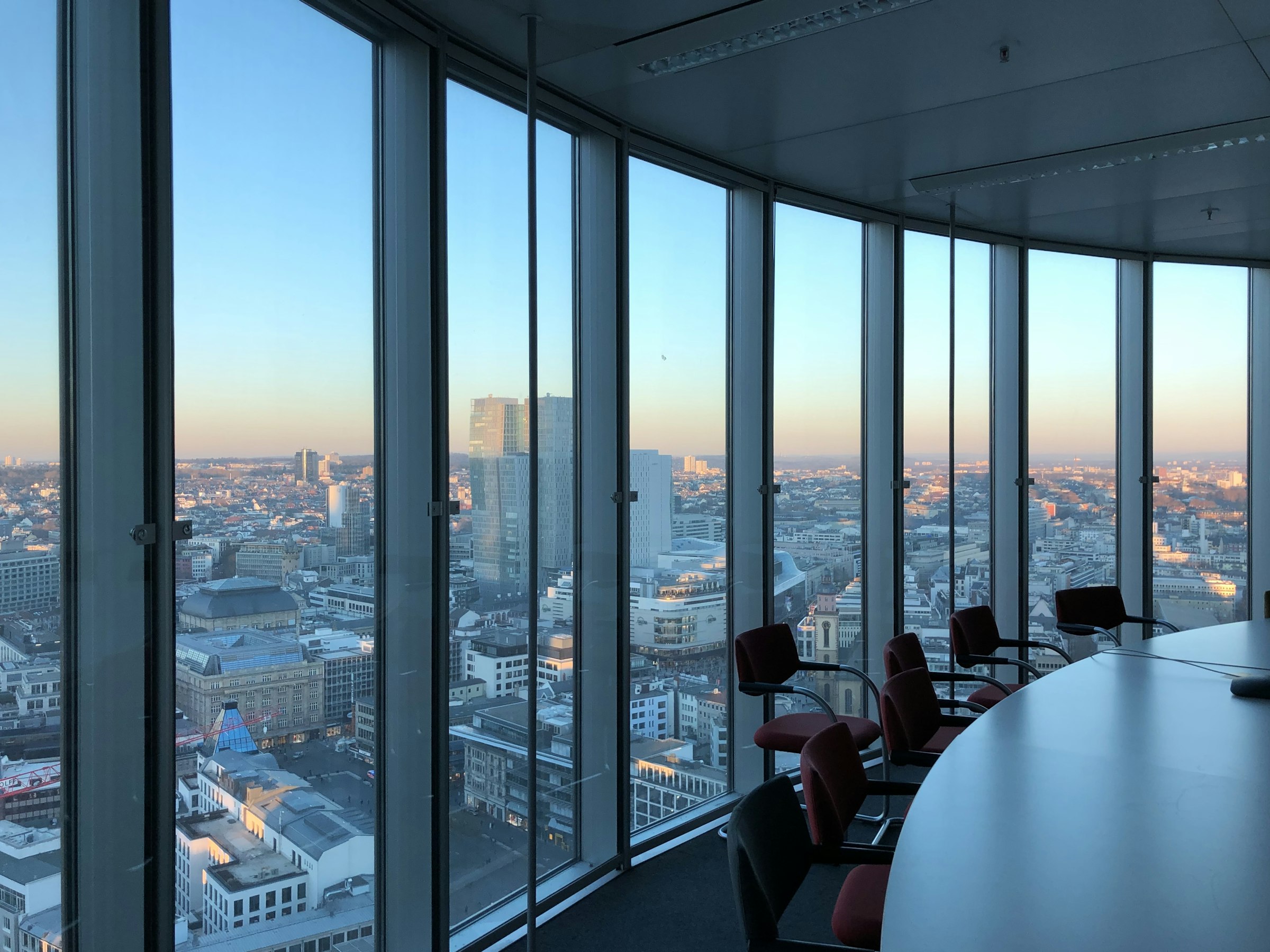 Empty boardroom behind floor-to-ceiling glass, high above a city at dusk